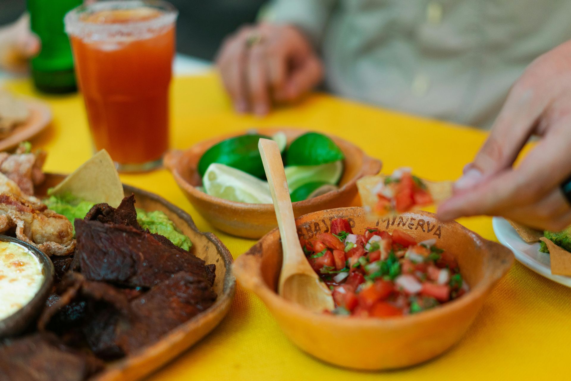 A spread of Mexican food on a yellow table, including a platter of meat, bowls of salsa and lime wedges, and a beverage.