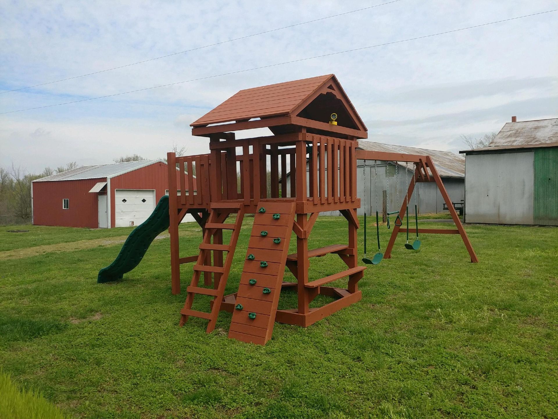 Brown wooden playset with a slide, climbing wall, swing set, and playhouse roof on green grass.