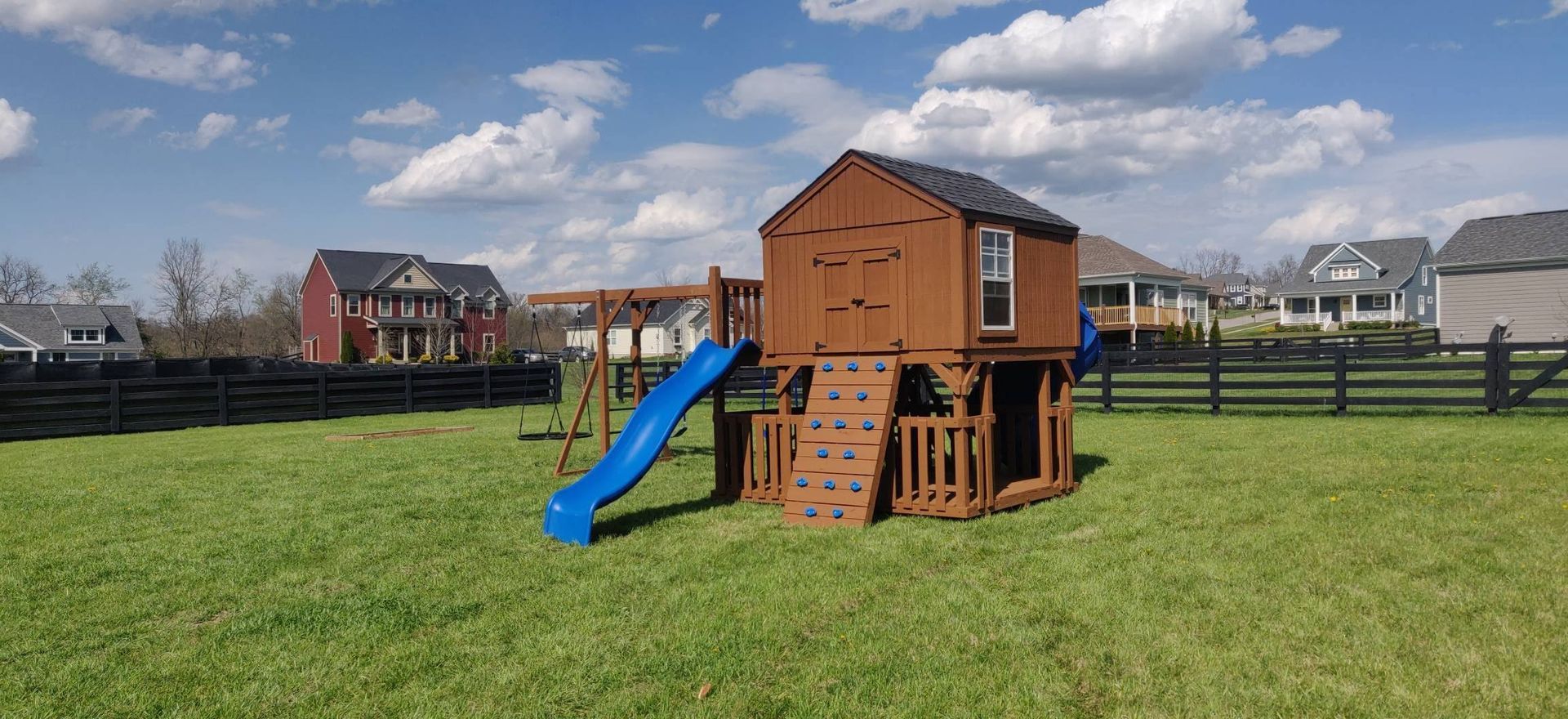 A brown wooden playset with a blue slide, climbing wall, and swing set in a grassy backyard, blue sky.