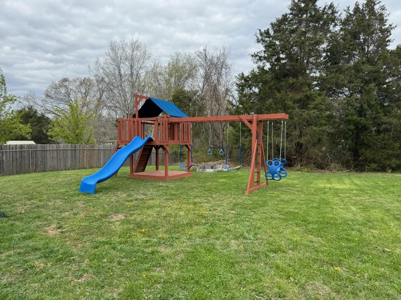 Wooden backyard playset with stairs, slide, and swing set on a grassy lawn under cloudy skies