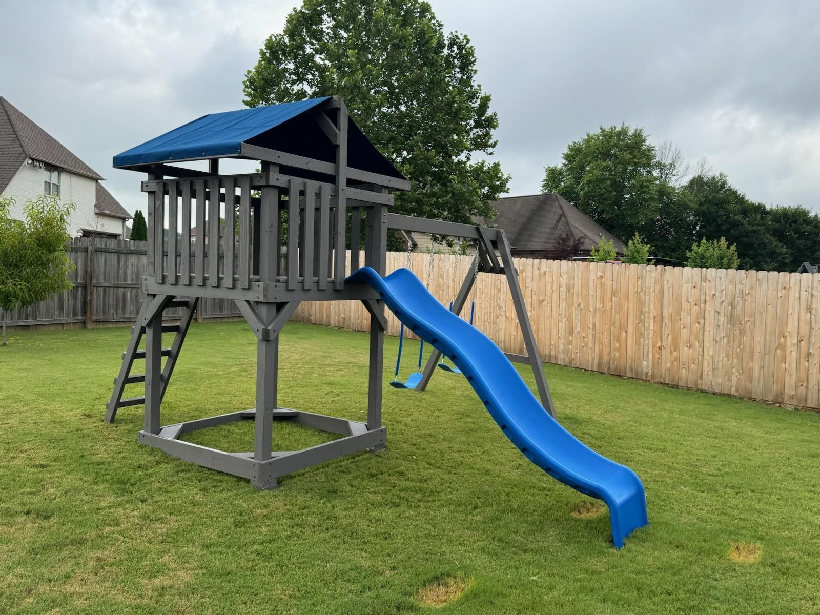 Playground with gray wooden structure, blue slide and swing set, on green lawn.