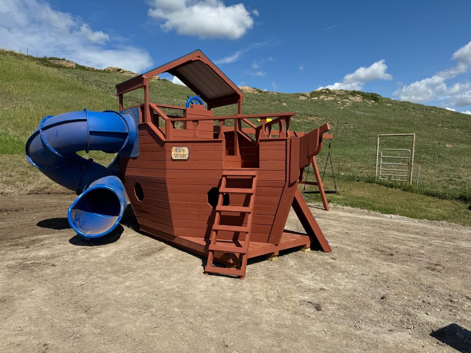 Playground with wooden ship structure, blue slide, brown ladder, on a gravel ground, with green hills and blue sky.