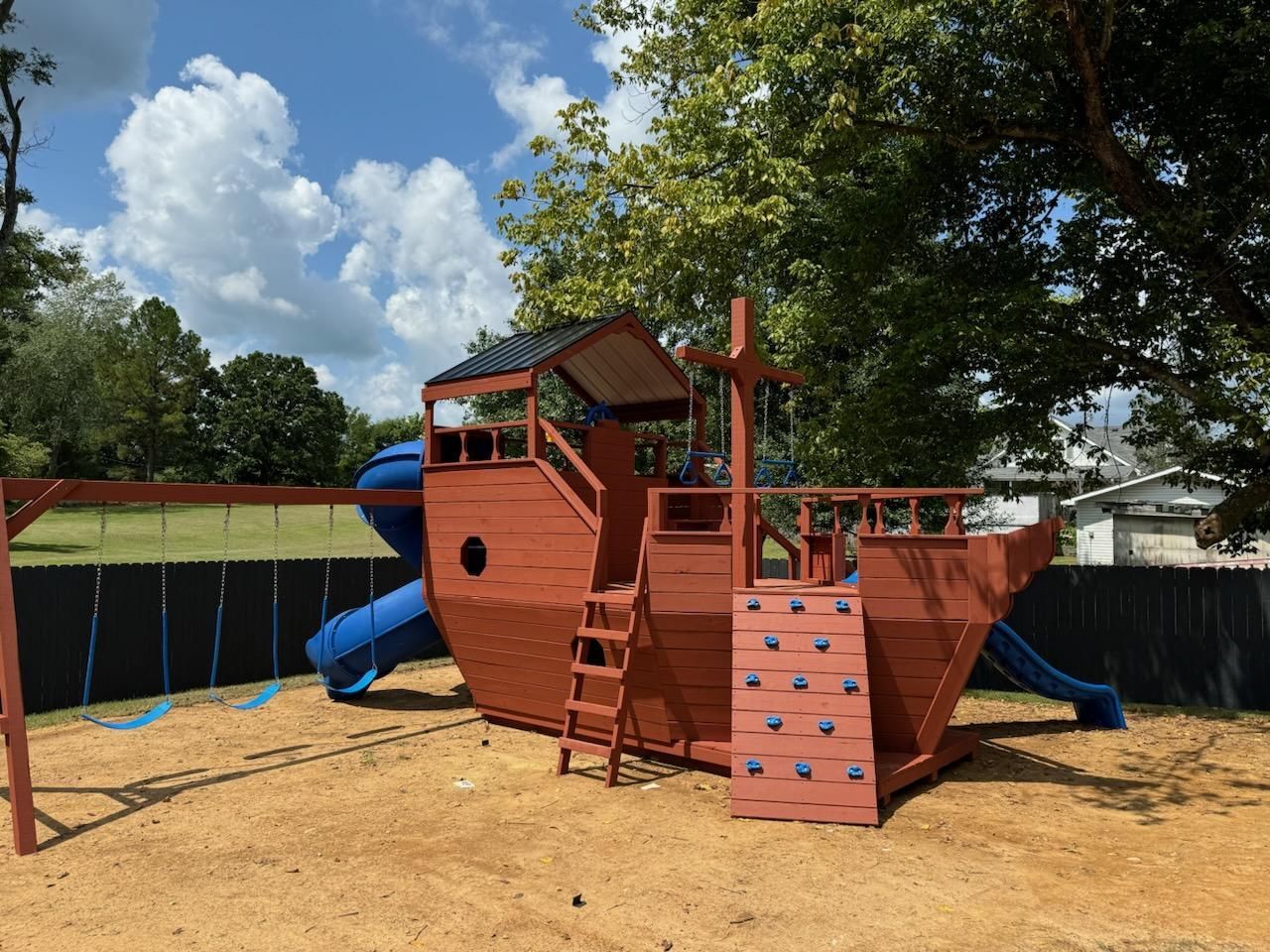 Playground with a wooden ship structure, blue slides, swing set, on a dirt ground, under a cloudy sky.