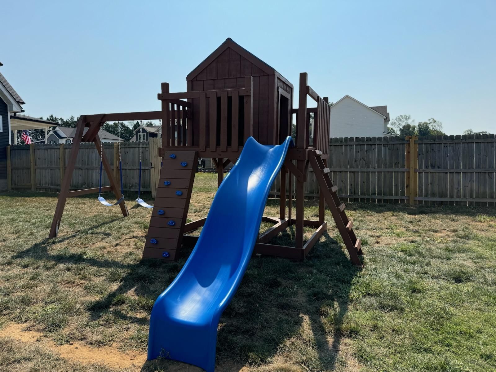 Wooden playset with blue slide, swings, and climbing wall in a yard.