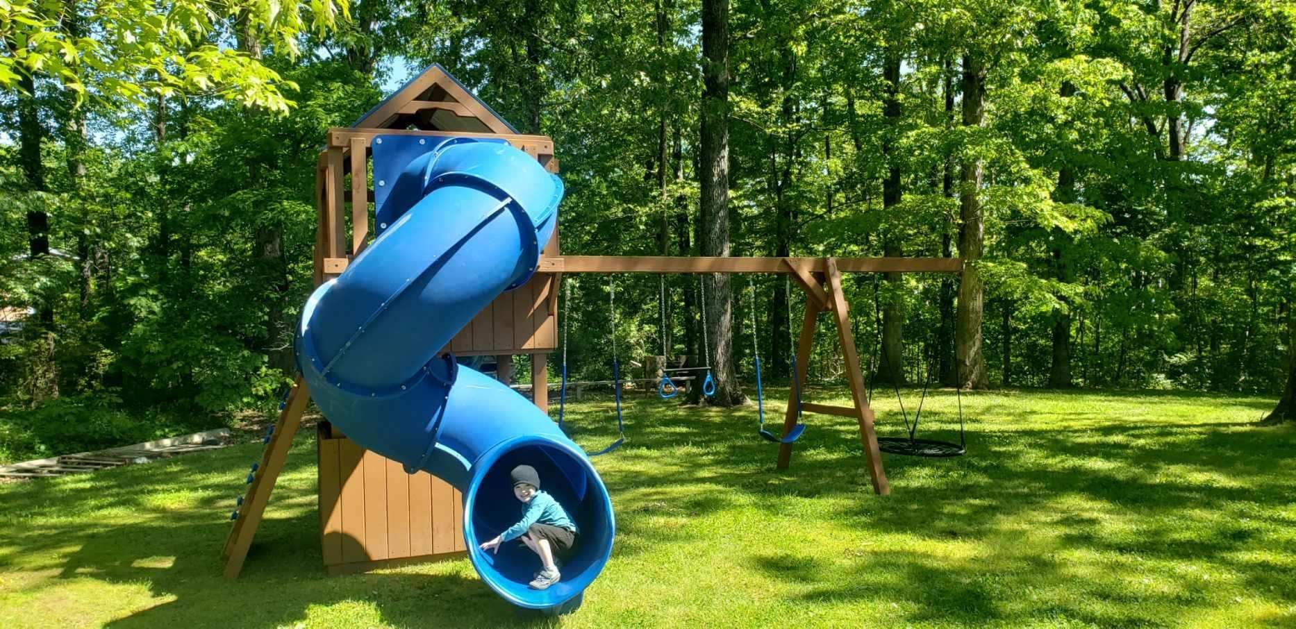 Child sliding down a blue spiral slide in a backyard with a swing set. Green grass and trees surround them.