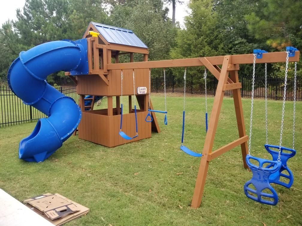 Brown wooden playset with blue tunnel slide and swings on green lawn.
