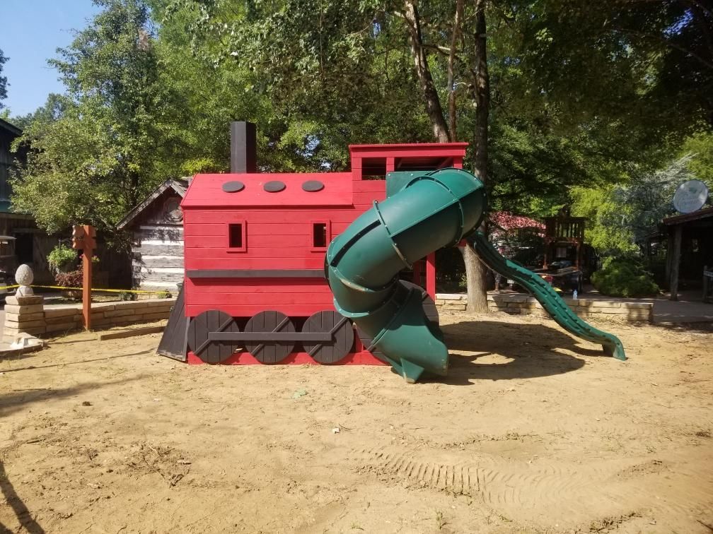 Red train-shaped playground with a green slide in a sandy area, outdoors.