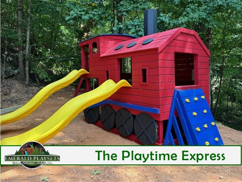 Red playhouse with yellow slides and blue climbing steps in a wooded playground.