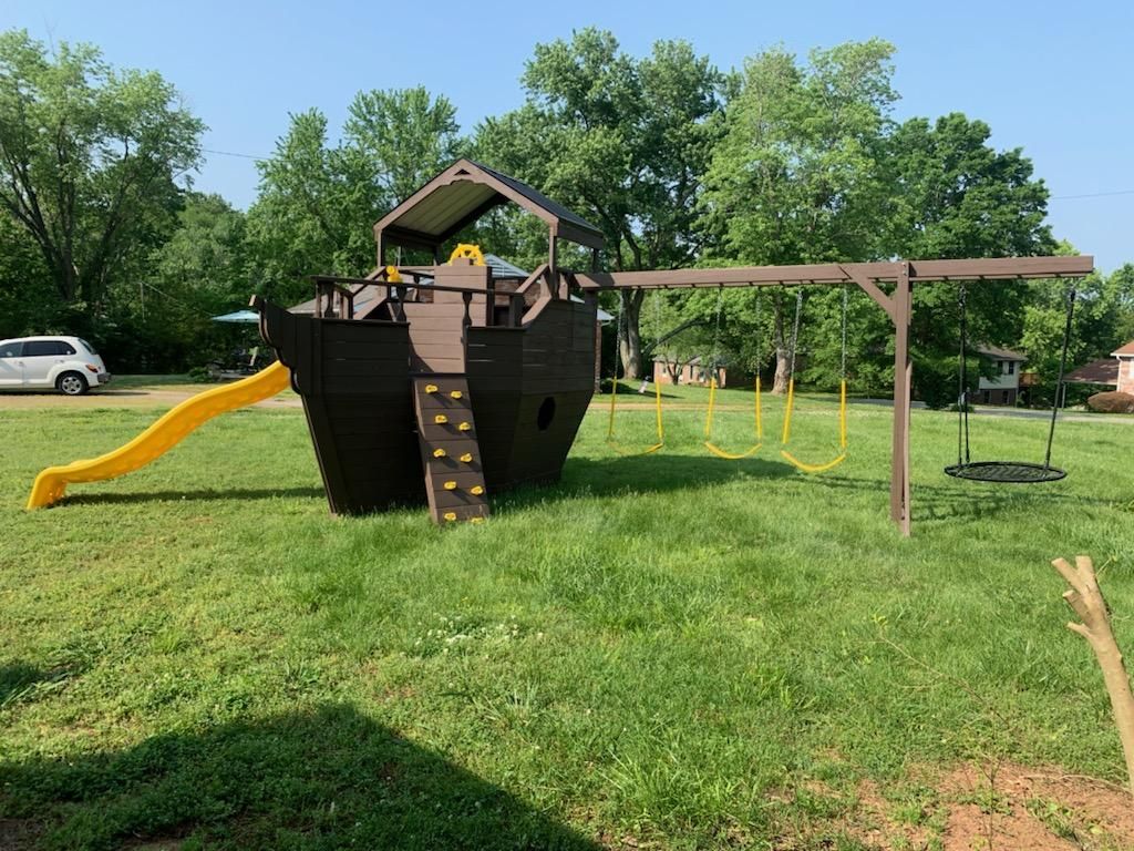 Play structure resembling a ship with a slide, climbing wall, and swingset in a grassy yard.