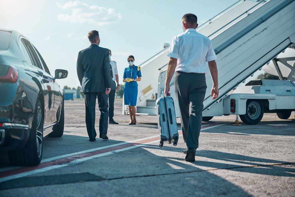 People boarding a plane: a man with a suitcase walks toward steps. Others walk in the background. Blue sky.