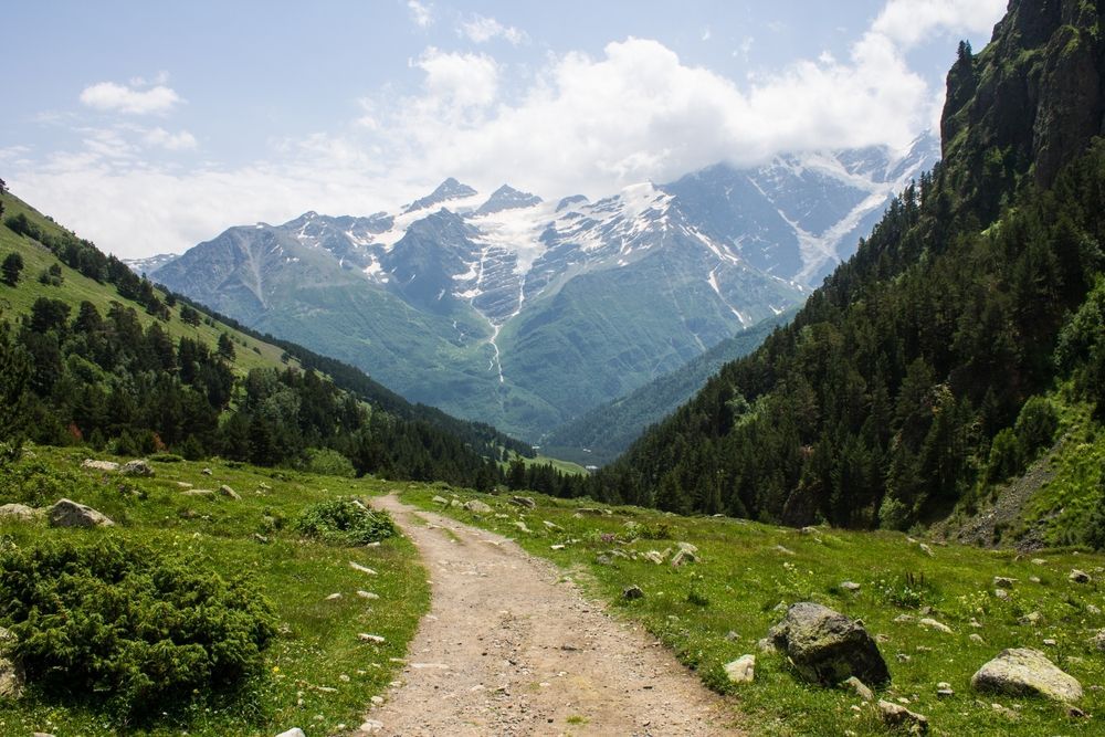 Dirt path through a green valley towards snow-capped mountains. Sunny with cloudy skies.