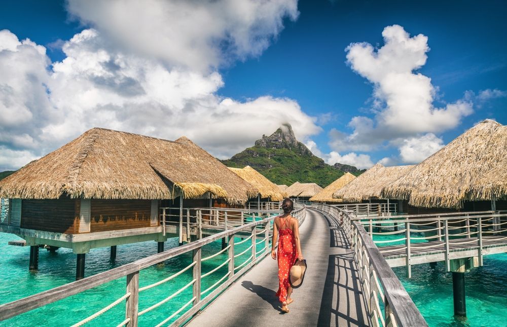 Woman in red dress walks on pier toward overwater bungalows, turquoise water, mountain background.