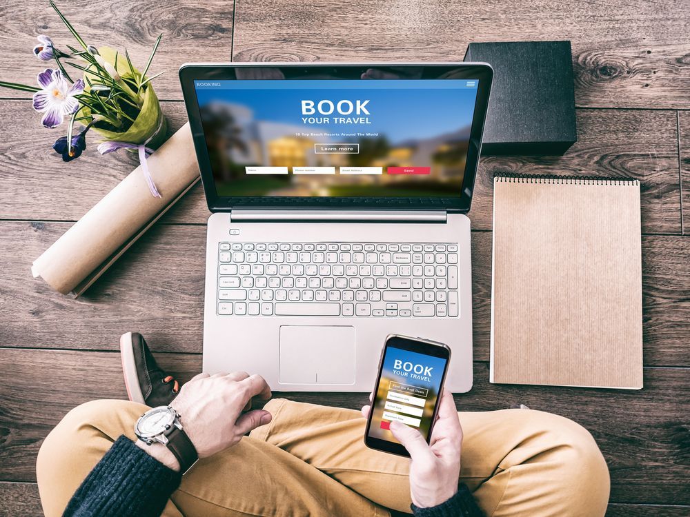 Person using laptop and smartphone to book travel; desk setup with notebook, flowers, and book.