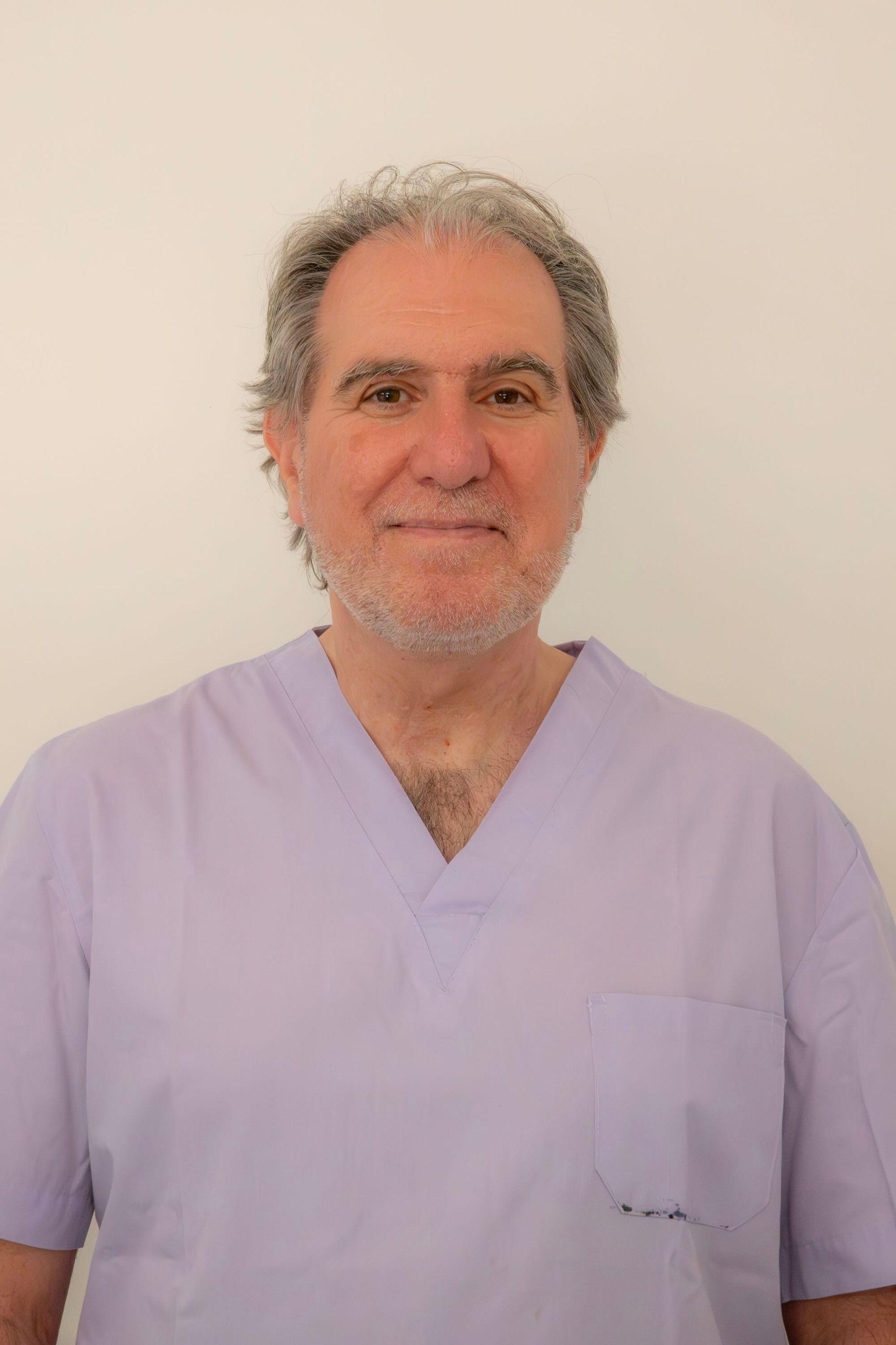 Un uomo con i capelli lunghi fino alle spalle e una leggera barba incolta, che indossa una casacca medica color lilla chiaro, sorride davanti a delle tende bianche.