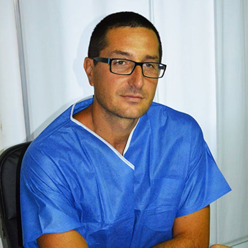 Una persona con gli occhiali e una casacca medica blu, guarda verso la telecamera su uno sfondo bianco uniforme.