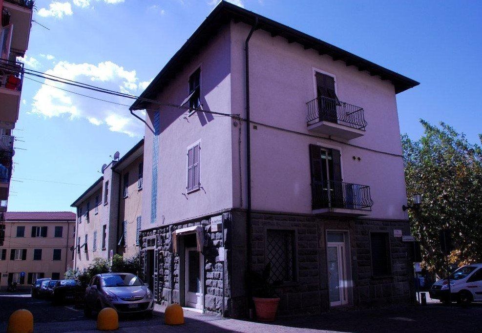 Un edificio di tre piani di colore chiaro, con fondamenta in pietra e balconi, situato all'angolo di una strada, con auto parcheggiate nelle vicinanze.