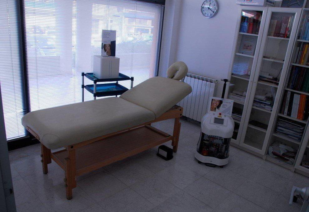 Una sala per trattamenti con un lettino da massaggio beige, un carrello con ruote contenente una scatola bianca e un'apparecchiatura clinica.