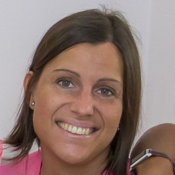 Una persona con i capelli castani lunghi fino alle spalle e una maglietta rosa sorride alla telecamera.