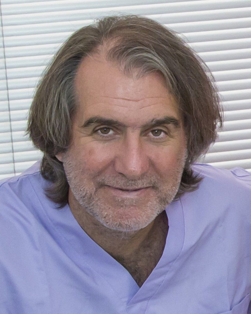 Un uomo con i capelli lunghi fino alle spalle e una leggera barba incolta, che indossa una casacca medica color lilla chiaro, sorride davanti a delle tende bianche.