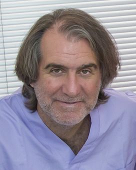 Un uomo con i capelli lunghi fino alle spalle e una leggera barba incolta, che indossa una casacca medica color lilla chiaro, sorride davanti a delle tende bianche.