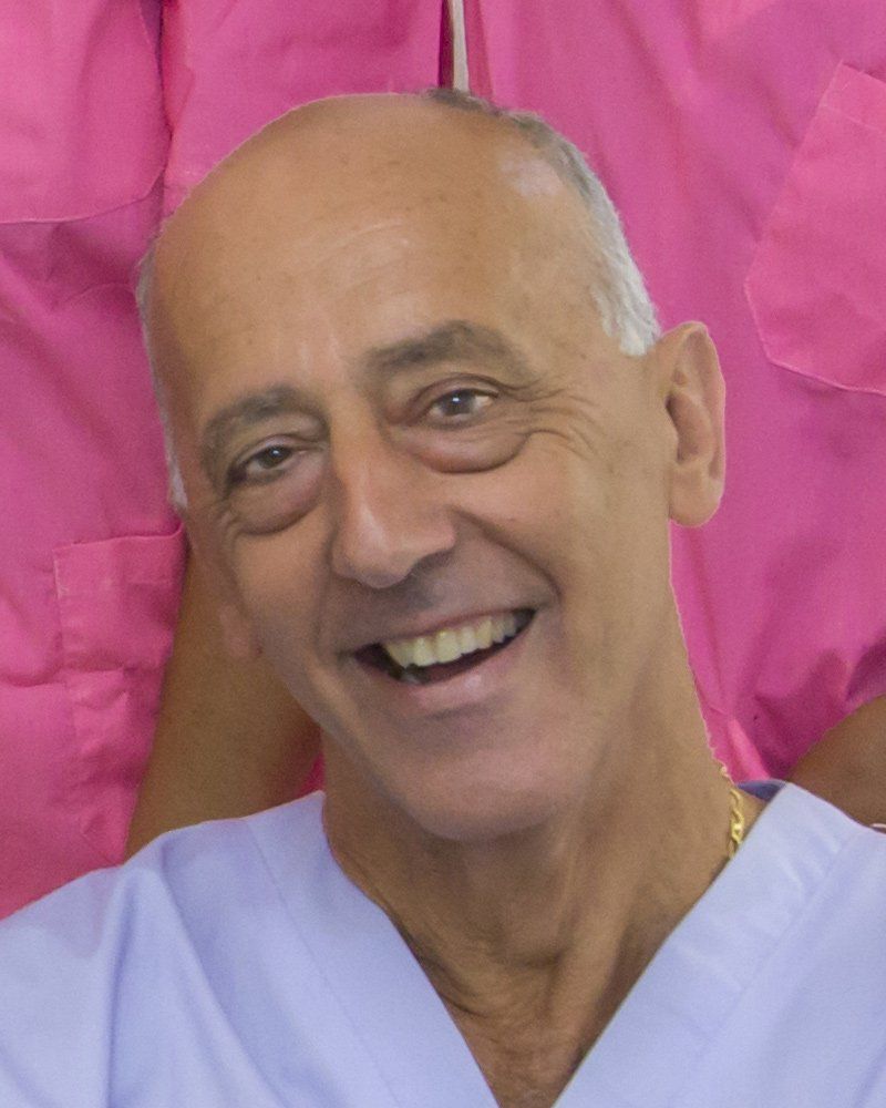 Ritratto ravvicinato di una persona sorridente che indossa una casacca chirurgica azzurra, su uno sfondo rosa sfocato.