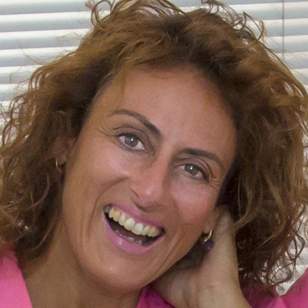 Una persona con i capelli castani ricci sorride ampiamente, indossando una maglietta rosa, sullo sfondo di tende a rullo orizzontali bianche.