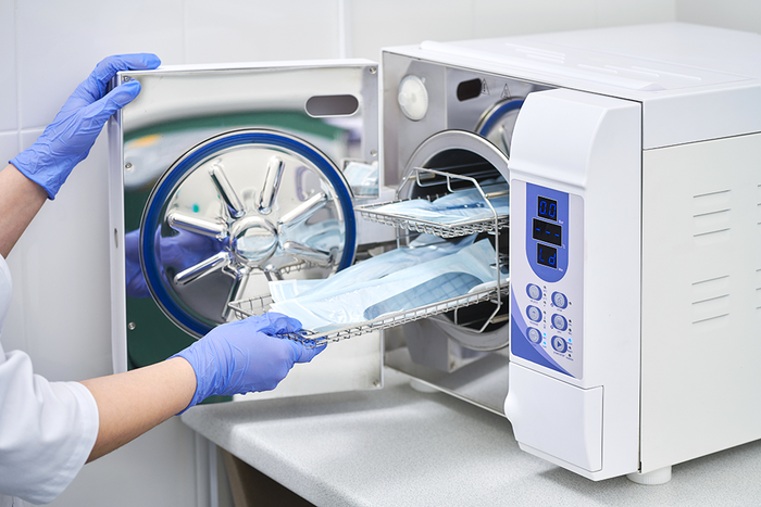 Una persona che indossa guanti medici blu fa scivolare un vassoio di strumenti sterili, avvolti in pellicola blu, all'interno di un'autoclave dentale aperta.