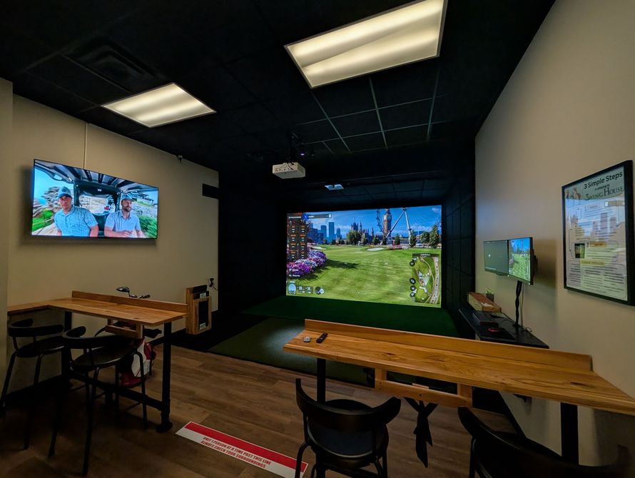 Golf simulator room: bar seating, screen displaying course, black ceiling, tan walls.