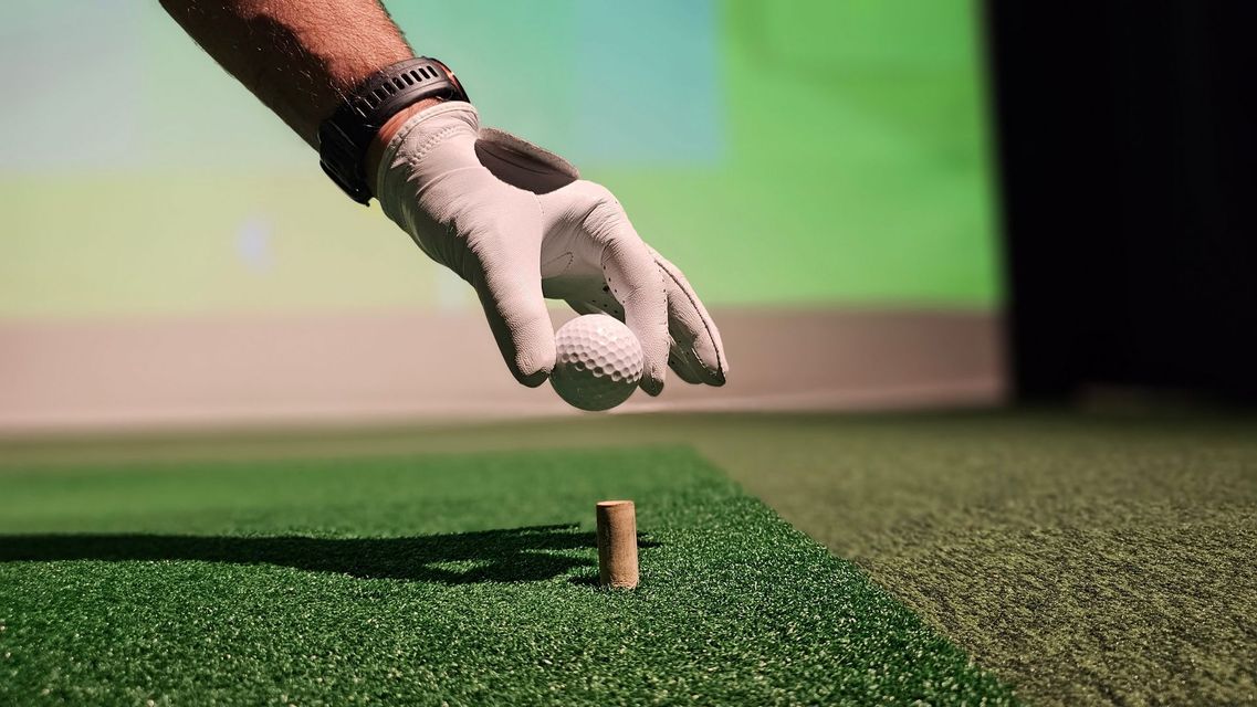 A golfer's gloved hand placing a golf ball on a tee on artificial turf, indoor setting.