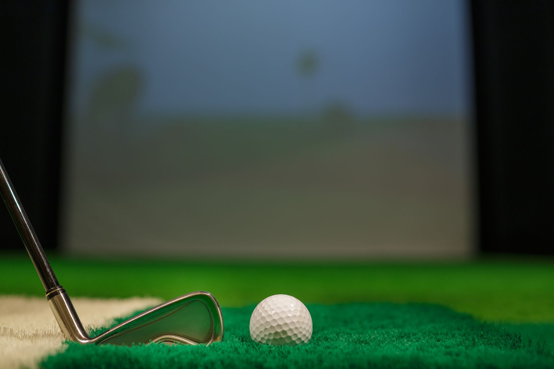 Golf club poised to hit a white ball on green turf, with a projection screen in the background.