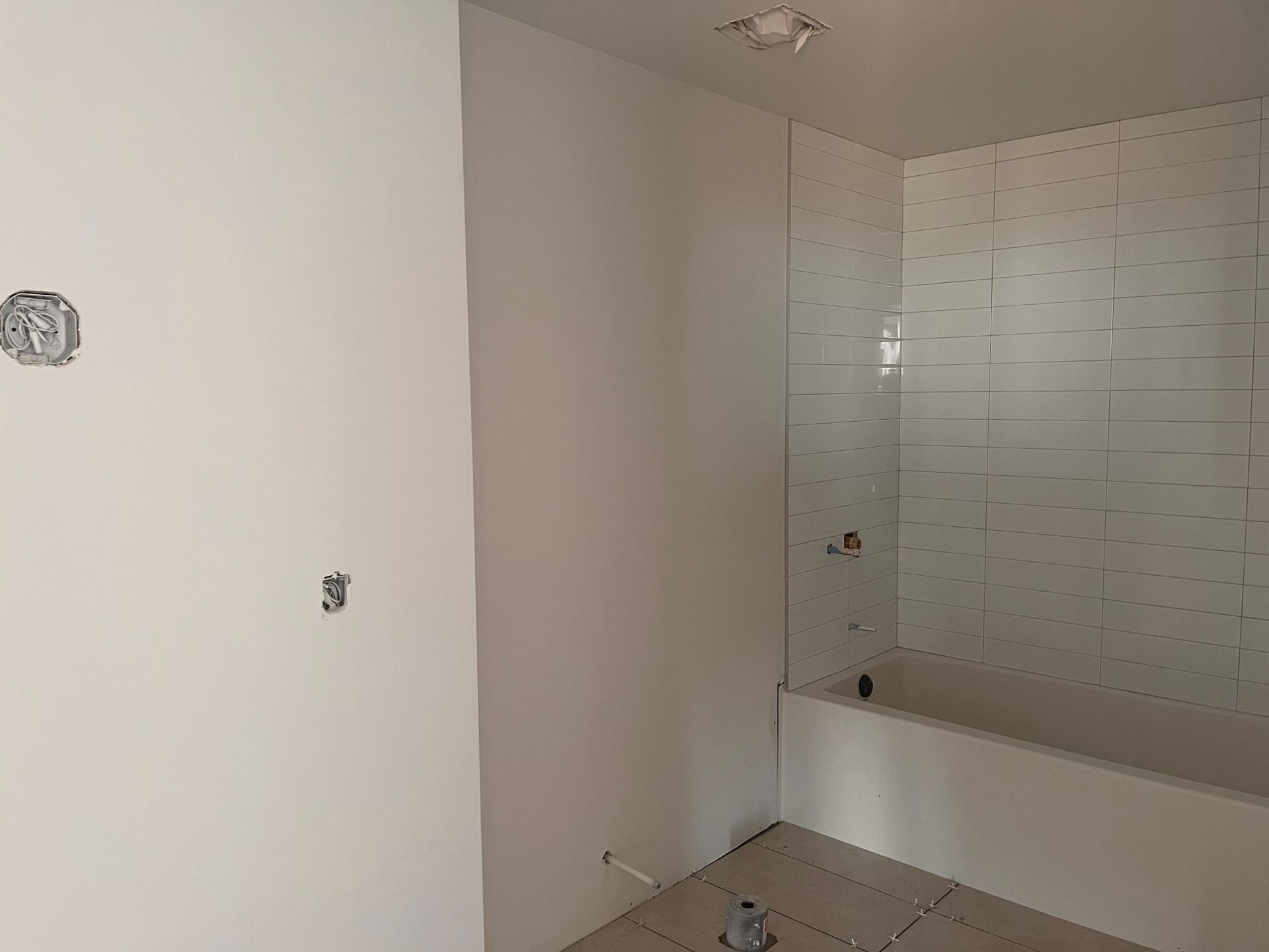 Salle de bains inachevée avec murs blancs, douche carrelée et baignoire blanche. Prises électriques visibles.