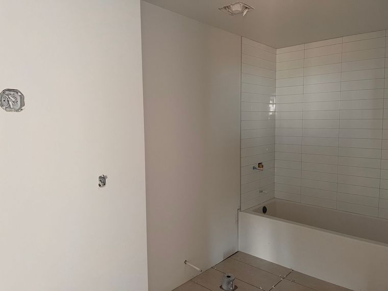 Salle de bains inachevée avec murs blancs, douche carrelée et baignoire blanche. Prises électriques visibles.