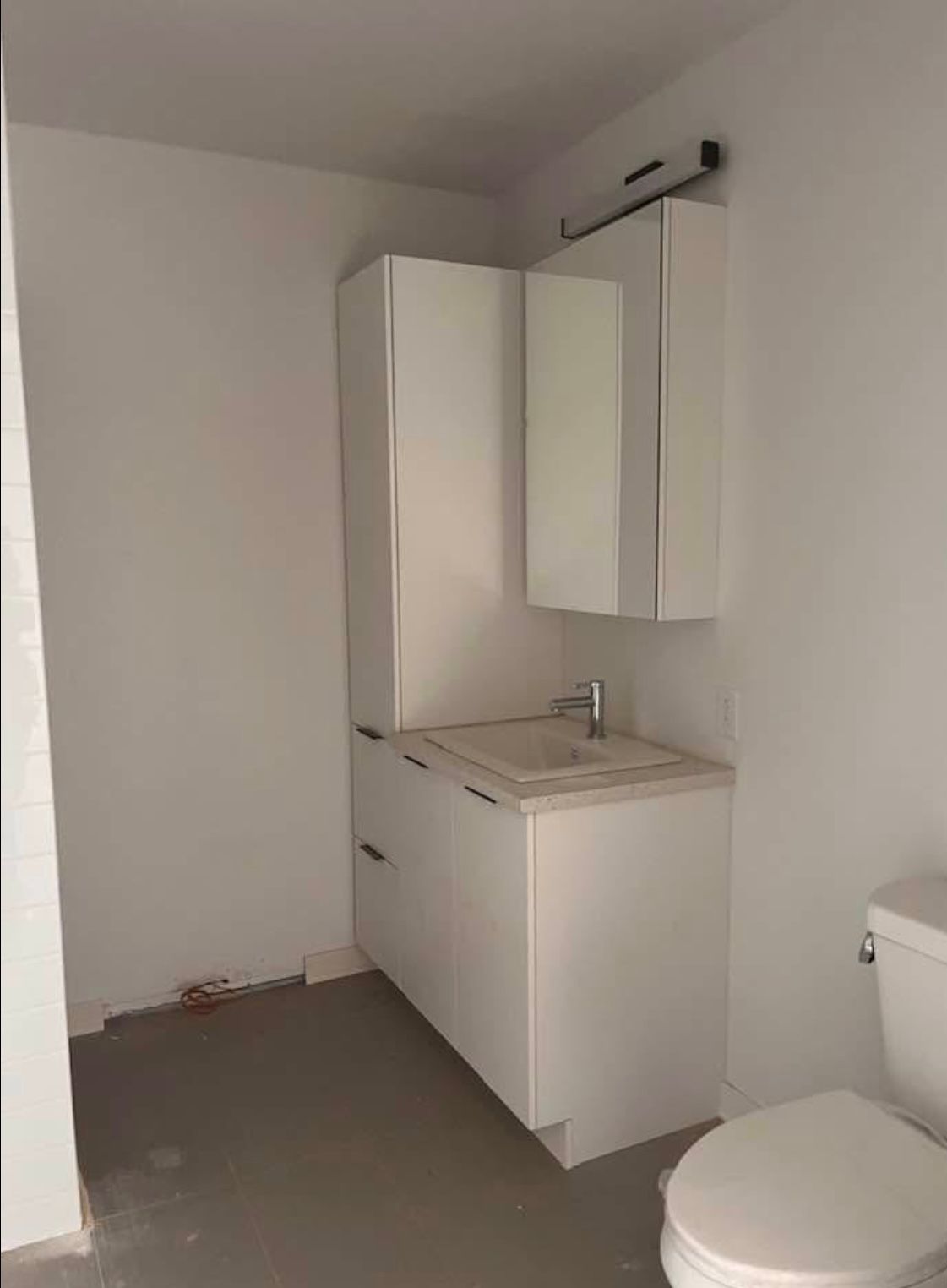 Salle de bains avec meubles blancs, lavabo et toilettes. Sol gris, murs blancs.