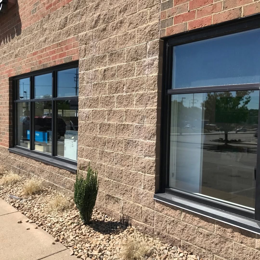Brick building exterior with black-framed windows and a sidewalk, reflected parking lot visible in glass