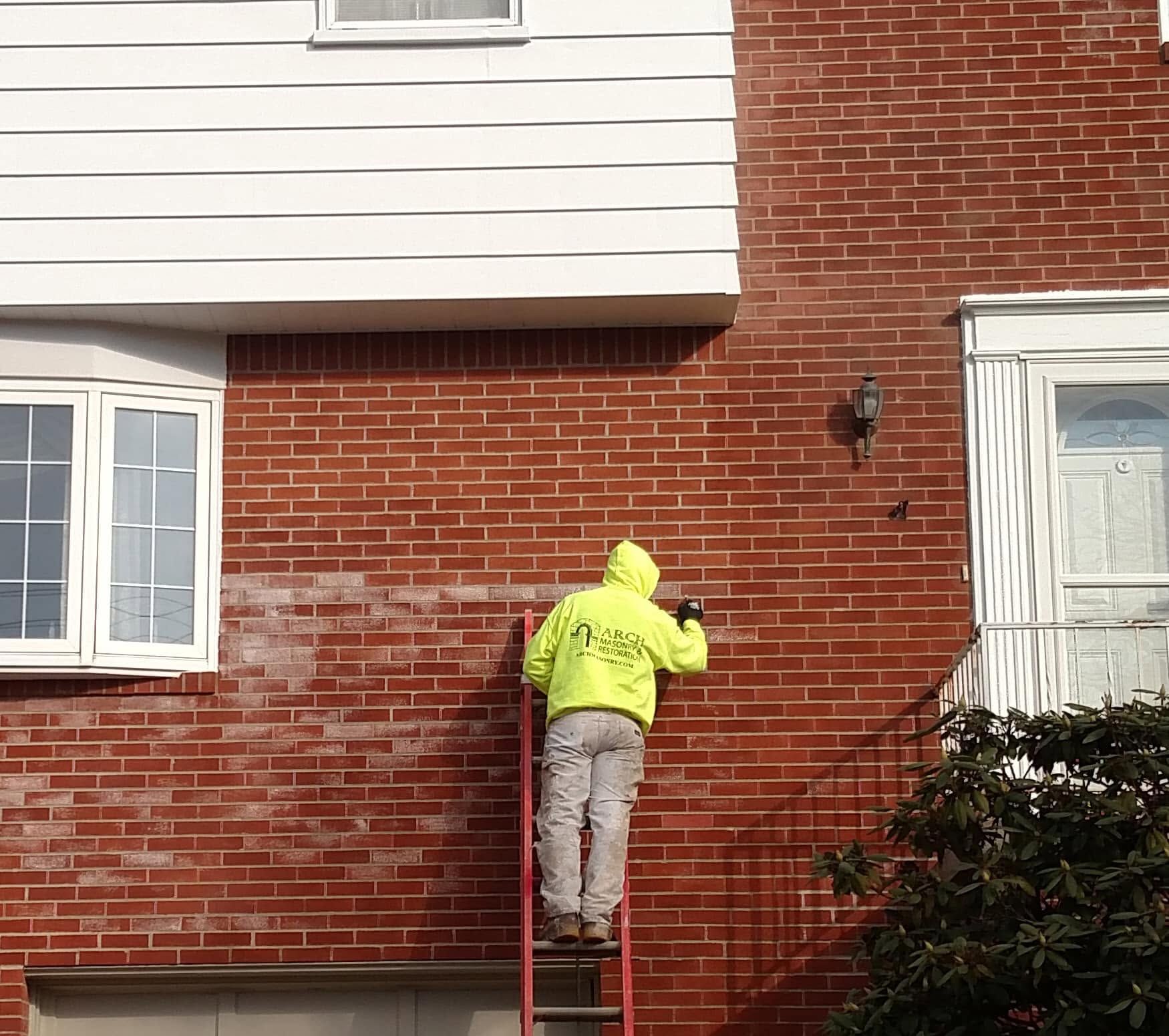 Person in a neon yellow jacket working on a brick house wall from a ladder