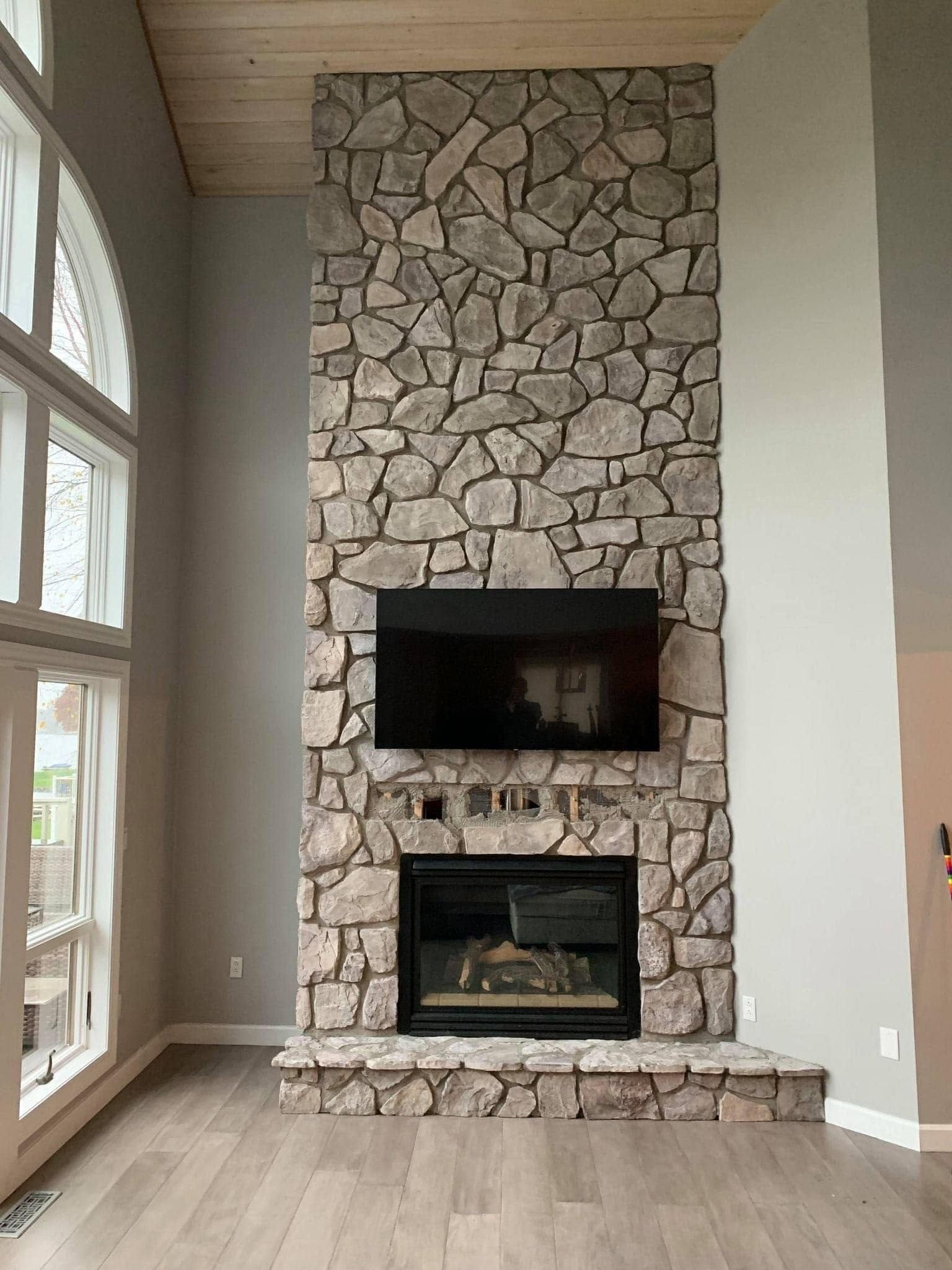 Stone fireplace with two black wall-mounted screens in a bright living room with tall windows