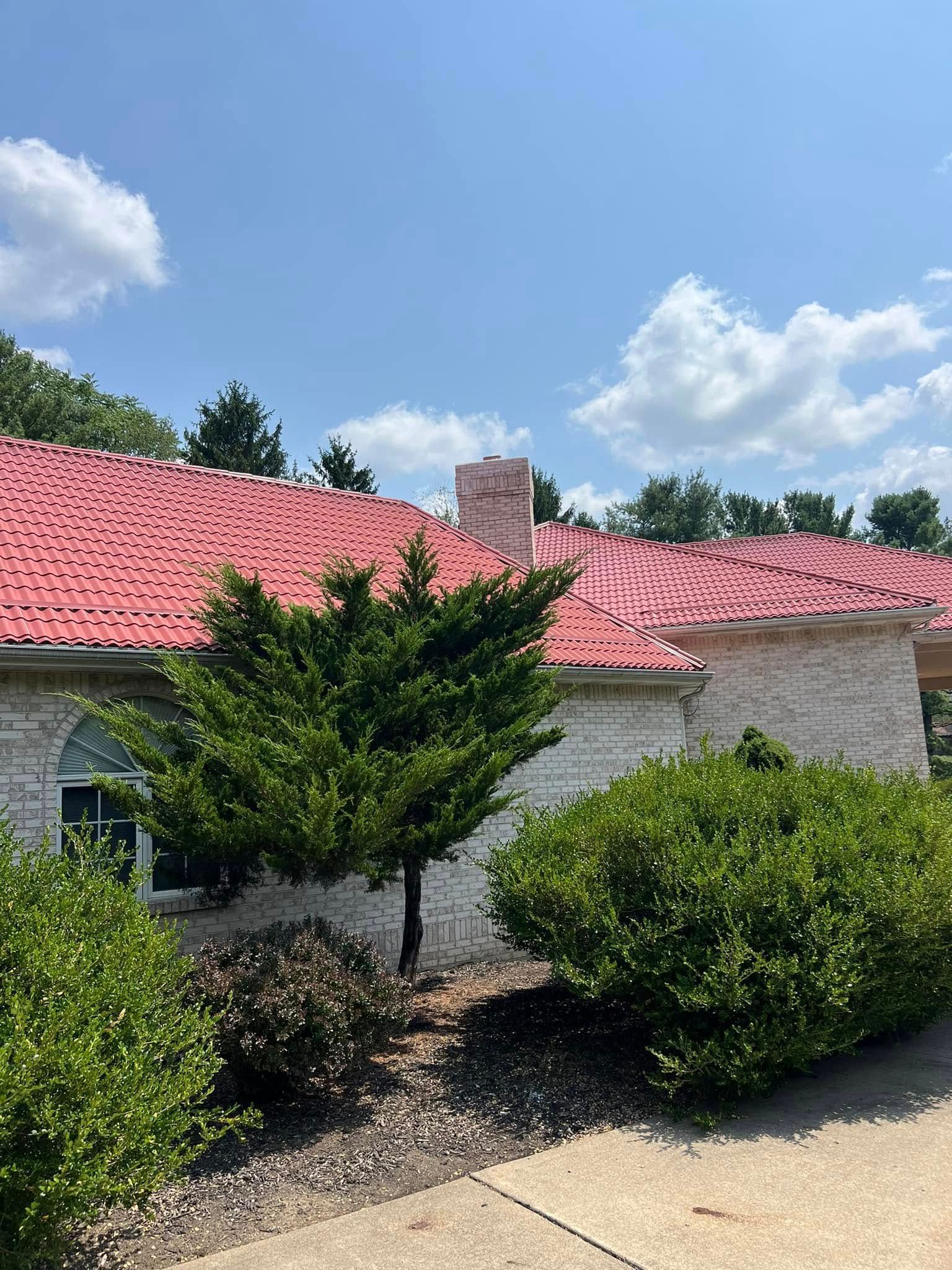 Single-story house with a red roof, white brick walls, and shrubs under a blue sky with clouds