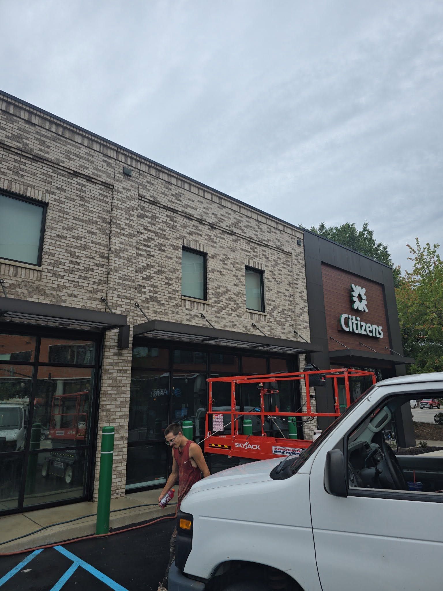 Brick storefront with glass doors, a red ladder lift, and a white van parked outside under cloudy skies