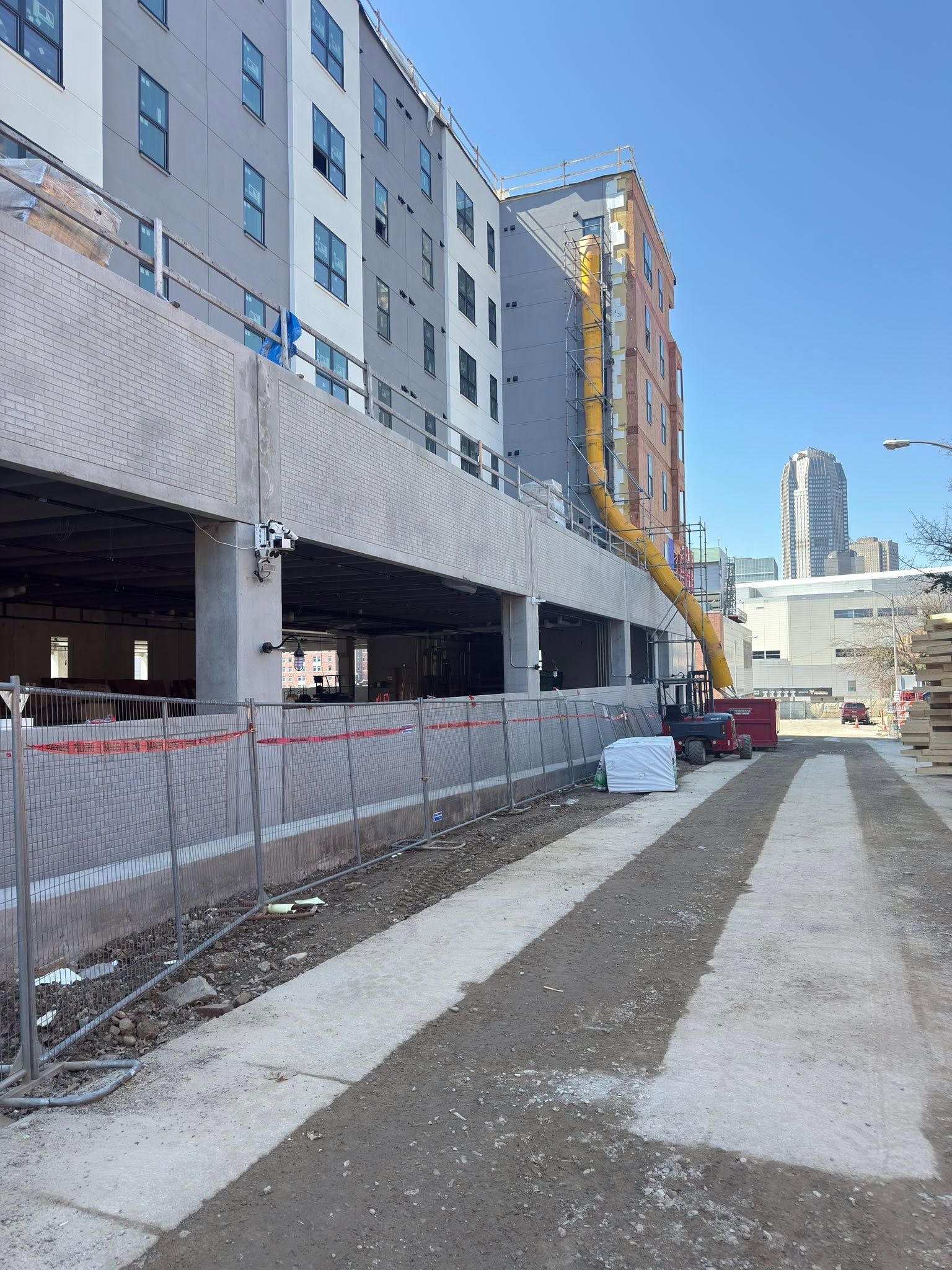 Construction site beside a multi-story building, with fencing, gravel path, and a yellow crane under a blue sky.