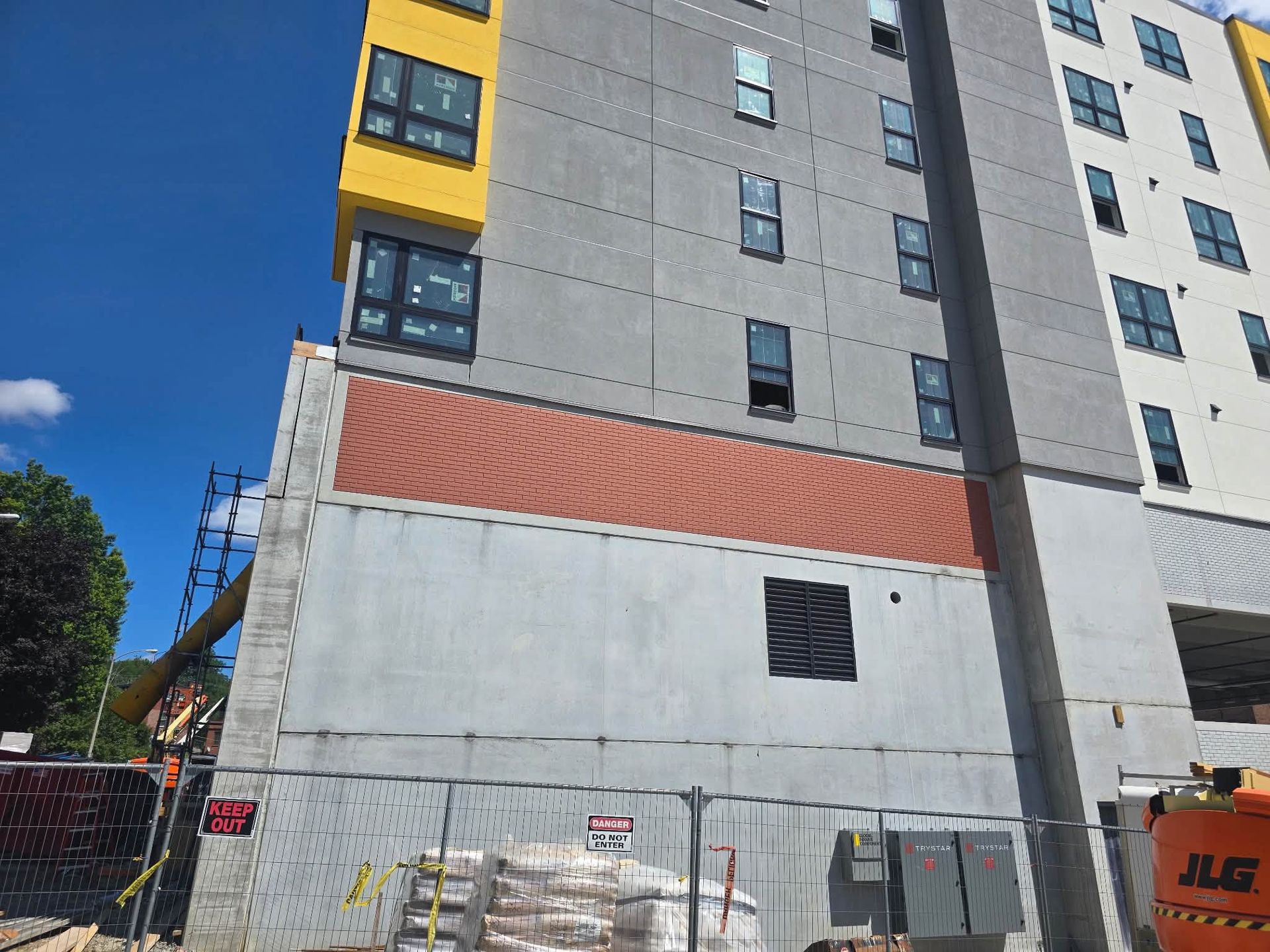 Multi-story building under construction with concrete walls, red paneling, and a fenced work area.
