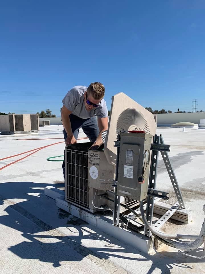 Technician inspecting rooftop HVAC unit under clear blue sky, with ladder and tools nearby.