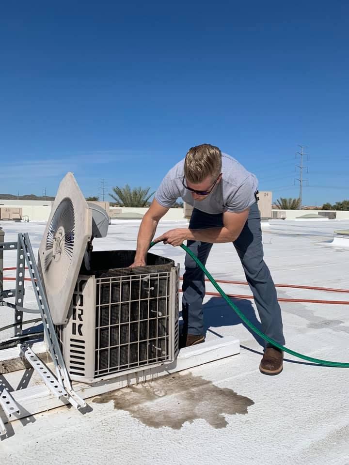 Person using a hose to wash an HVAC unit on a rooftop under a clear blue sky