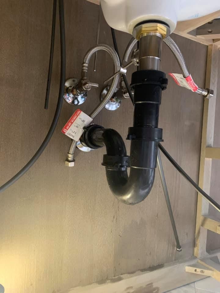 Under-sink plumbing with black drain trap, pipes, hoses, and shutoff valve on a wall-mounted sink.