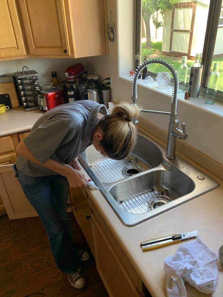 Person bends over a kitchen sink, rinsing dishes in a sunlit kitchen.