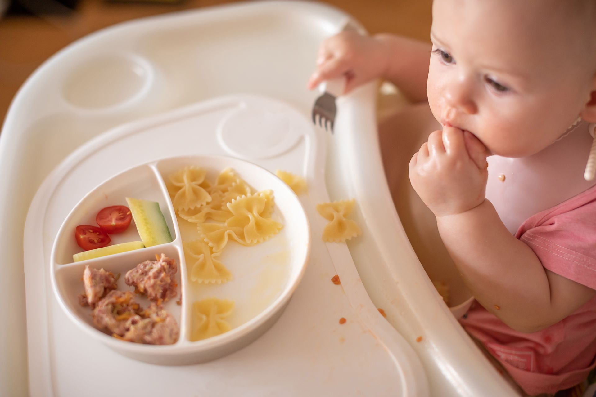 Bebé en silla alta comiendo pasta, tomates y carne de un plato dividido; utilizando un tenedor.