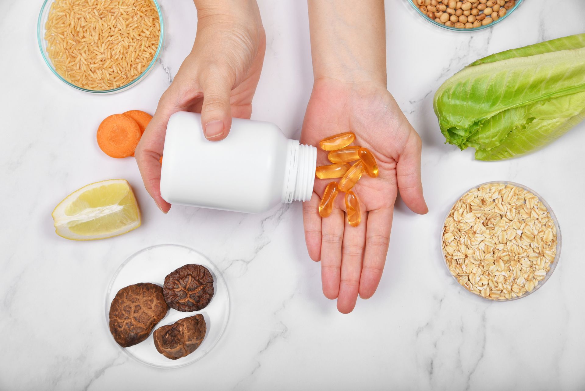 Manos vertiendo pastillas de una botella blanca, rodeada de alimentos como verduras y cereales.