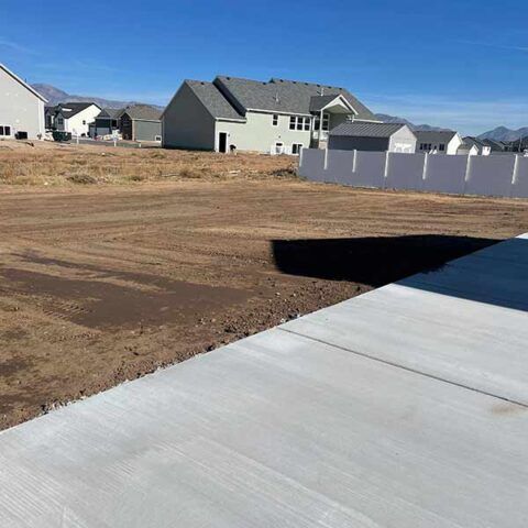 New concrete sidewalk beside a dirt lot and houses under a clear blue sky