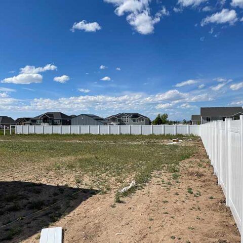 Empty dirt lot beside white vinyl fence under a blue sky with scattered clouds