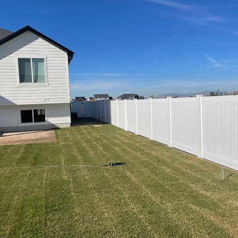 Backyard with green lawn, white vinyl fence, and a white two-story house under a clear blue sky