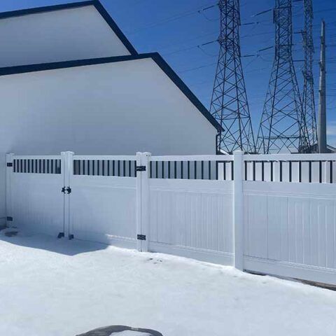 Snow-covered yard with a white fence and house wall under a clear blue sky, with power towers in the background